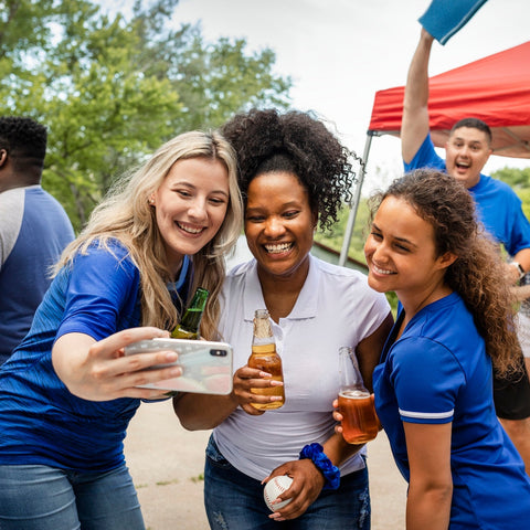 Three women taking a selfie at a outdoor event with drinks and a grill in the background.