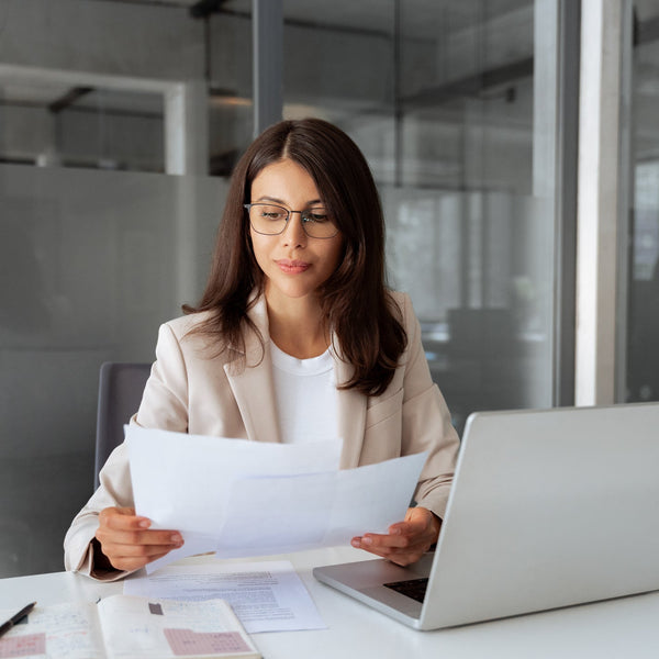 Woman in an office setting holding papers and using a laptop.