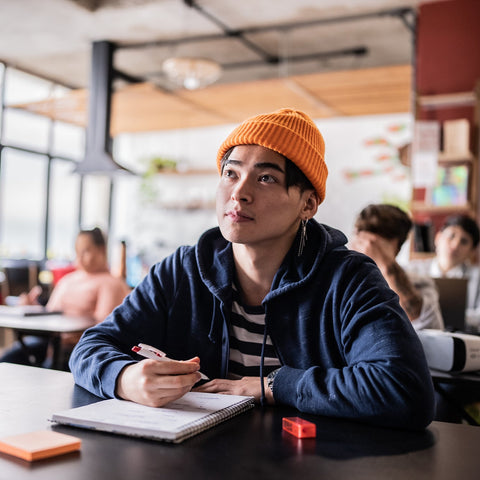 Male student wearing an orange beanie sitting at a table in a classroom setting.