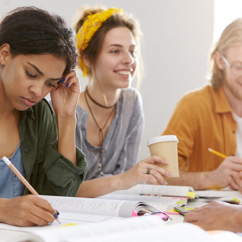 Three women sitting together, one writing in a notebook, another holding a coffee cup, and the third using a yellow pencil.