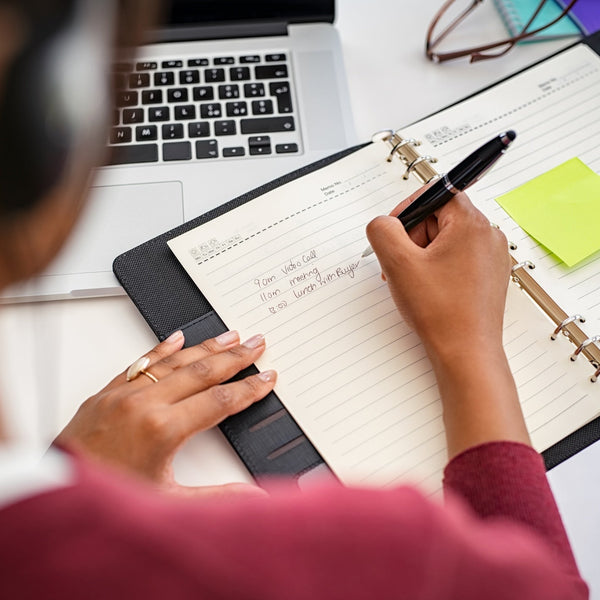 Person taking notes in a planner with a laptop and sticky notes on a desk.