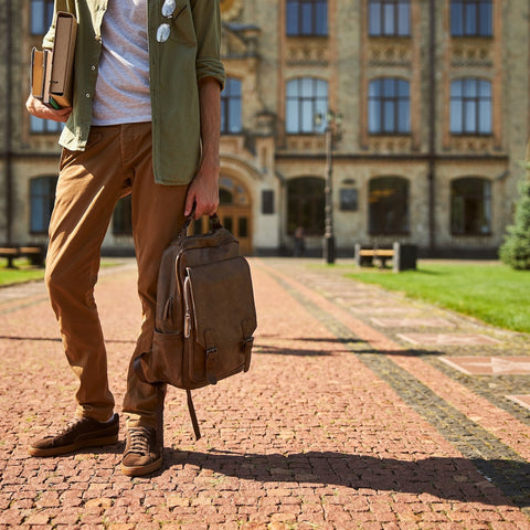 Person holding a brown backpack in front of a large building with a cobblestone path.